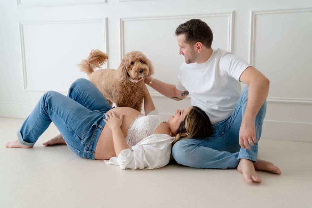 Pregnant woman lying on floor with man and fluffy dog in bright white studio maternity photo session.