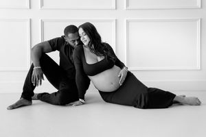 Black and white maternity photo of couple in relaxed pose on white floor against minimalist wall background.