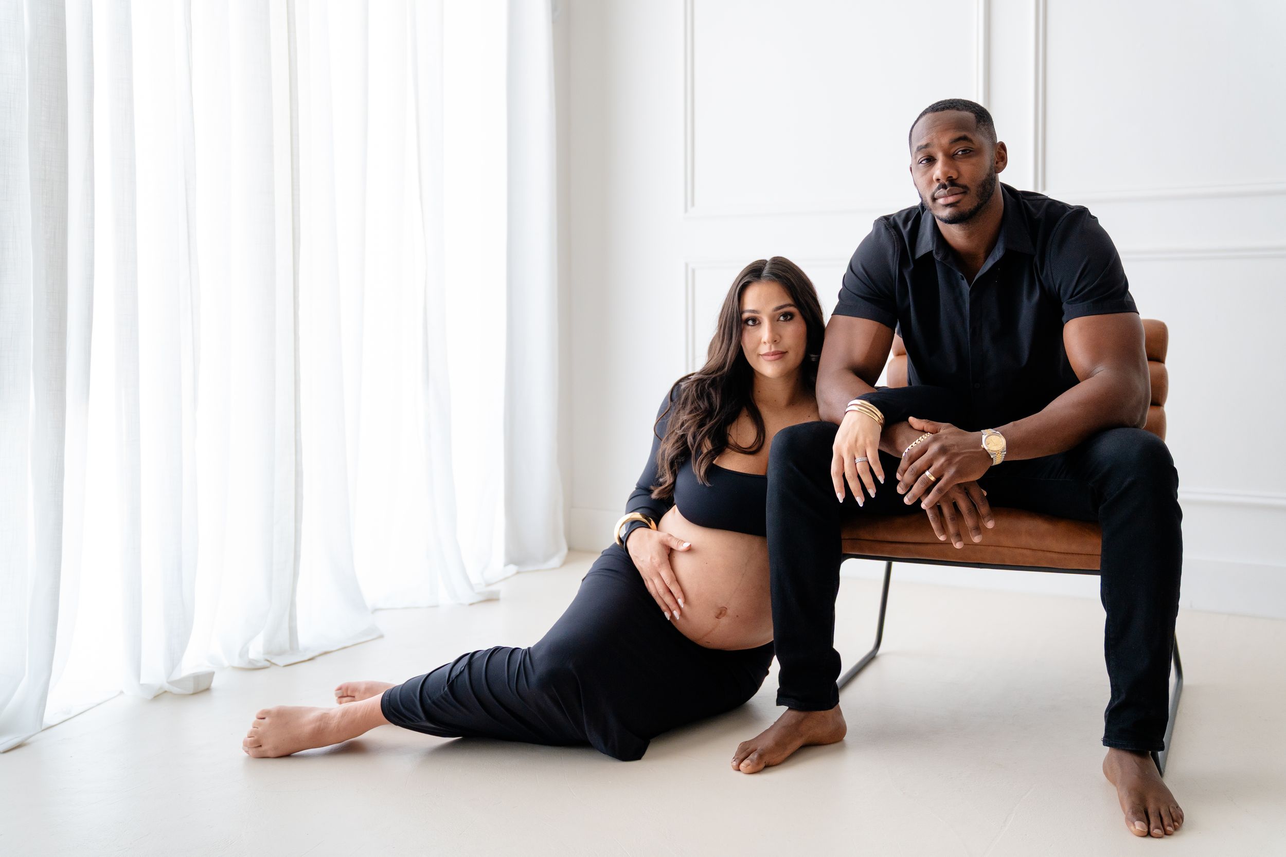 Two people wearing black outfits pose casually on a bright white floor against a white backdrop.