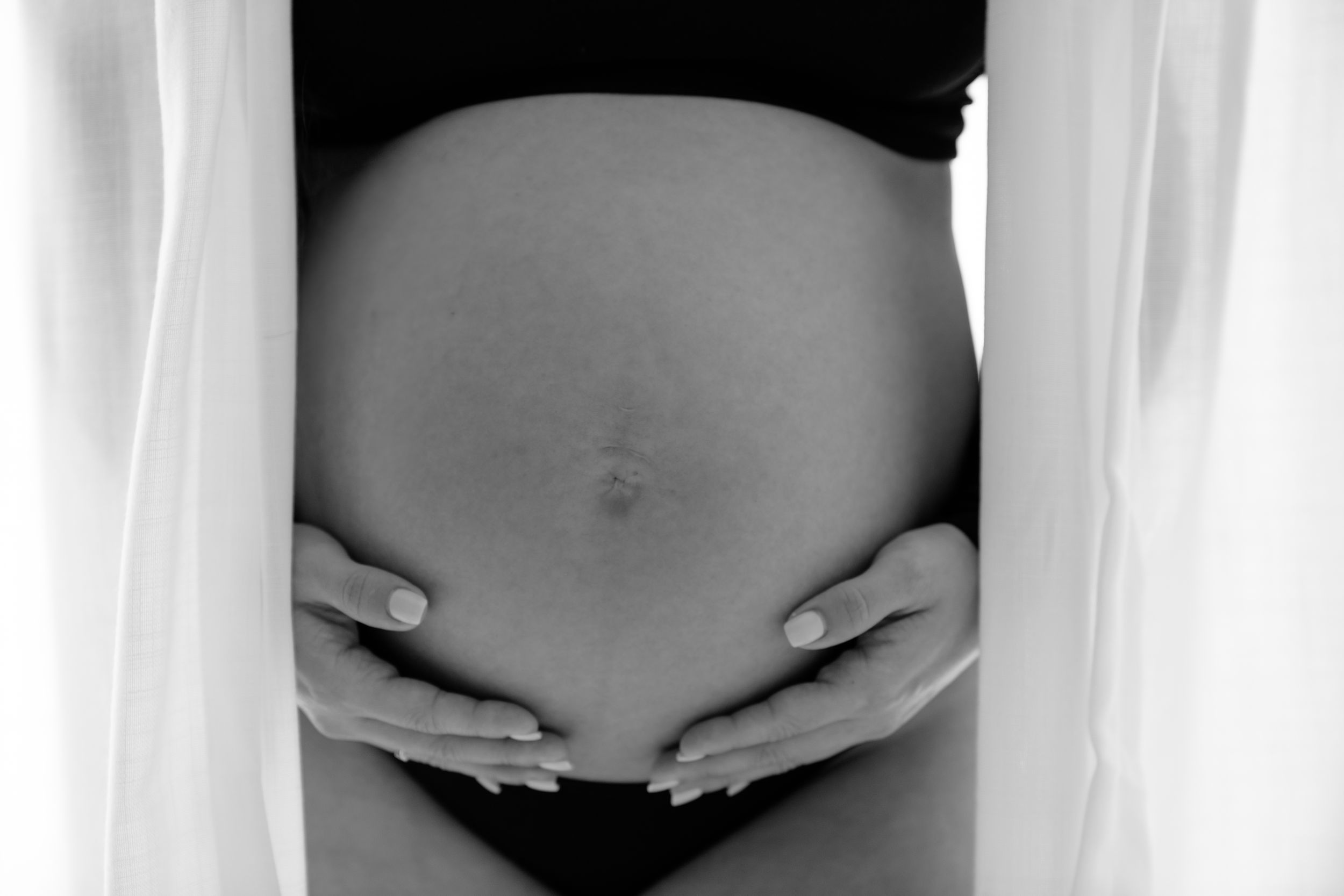 Black and white artistic photo of a pregnant belly being cradled by hands against a light fabric backdrop.