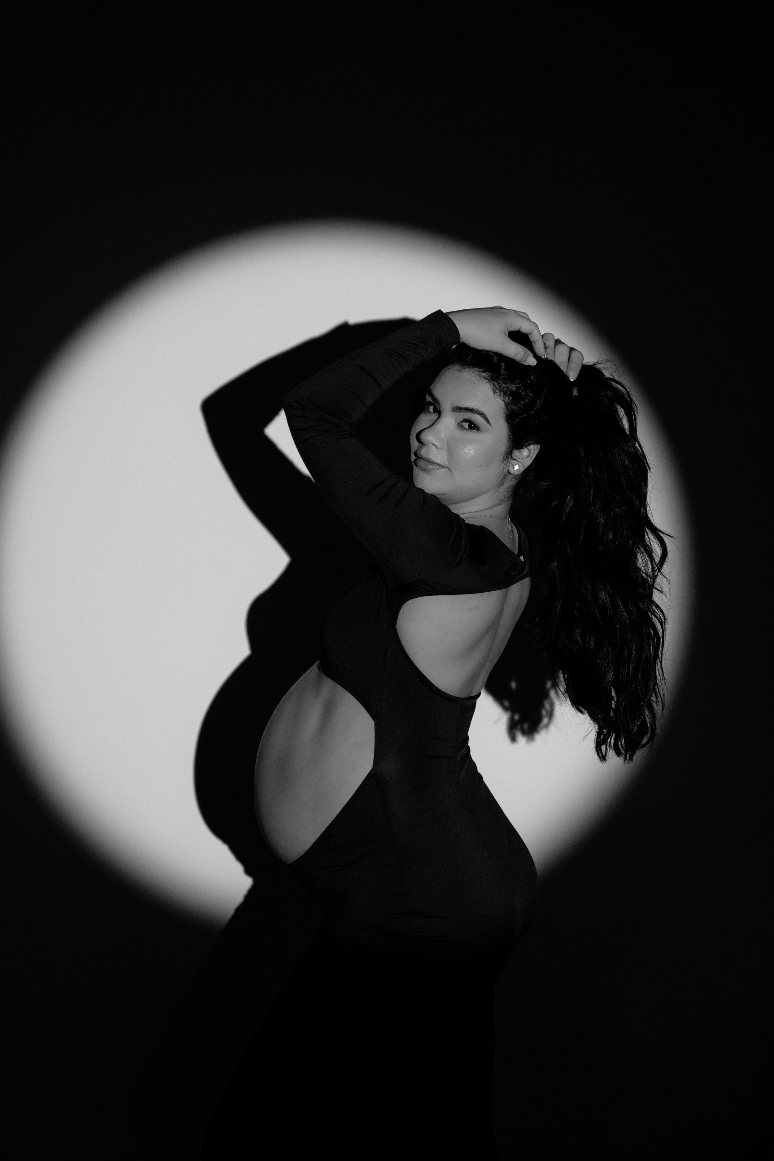 Black and white photo of a pregnant woman in a backless dress posing gracefully under a spotlight.