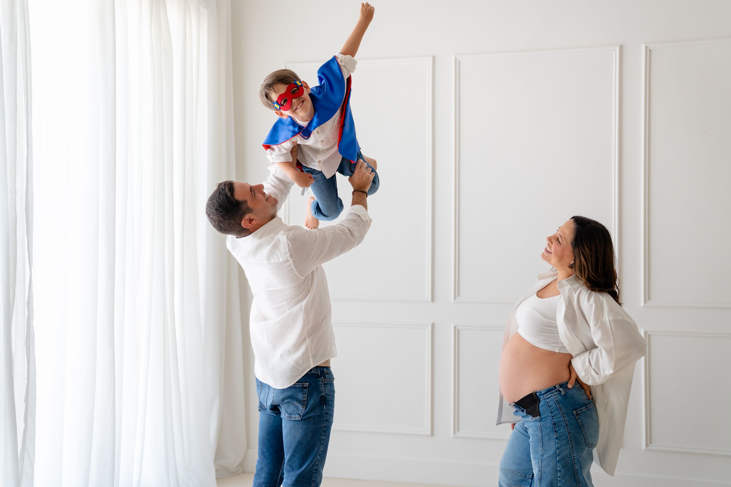 Pregnant woman smiles as man lifts child in superhero costume in bright white room during family photo session.