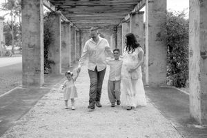 Black and white family photo of pregnant mother, father, and two children walking under a pergola outdoors.