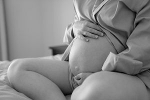A pregnant woman in white dress sits on bed and touches her belly while soft sunlight illuminates the room.