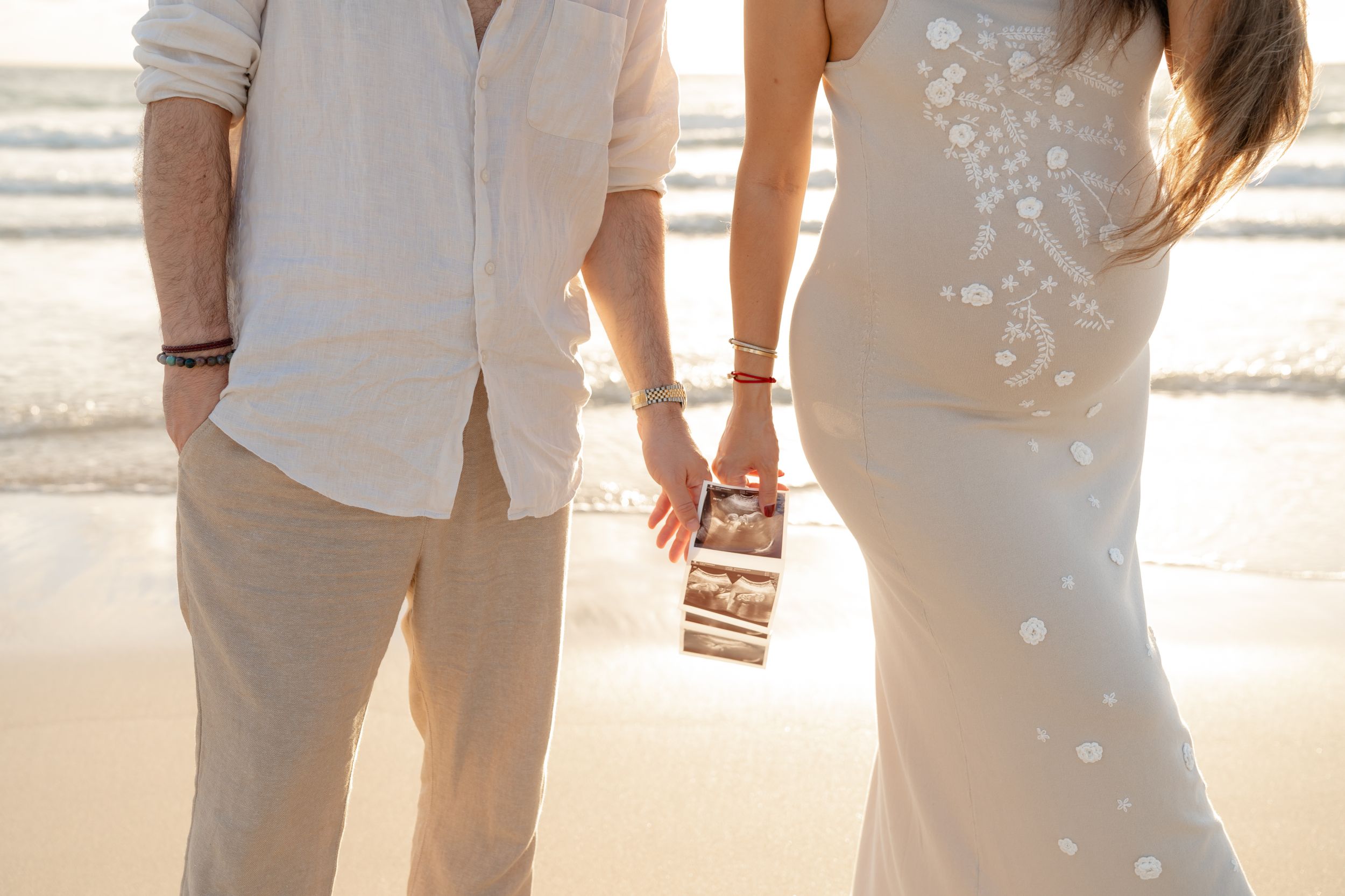 Couple holding hands on beach at sunset, woman in floral dress holding ultrasound photo, pregnancy announcement.