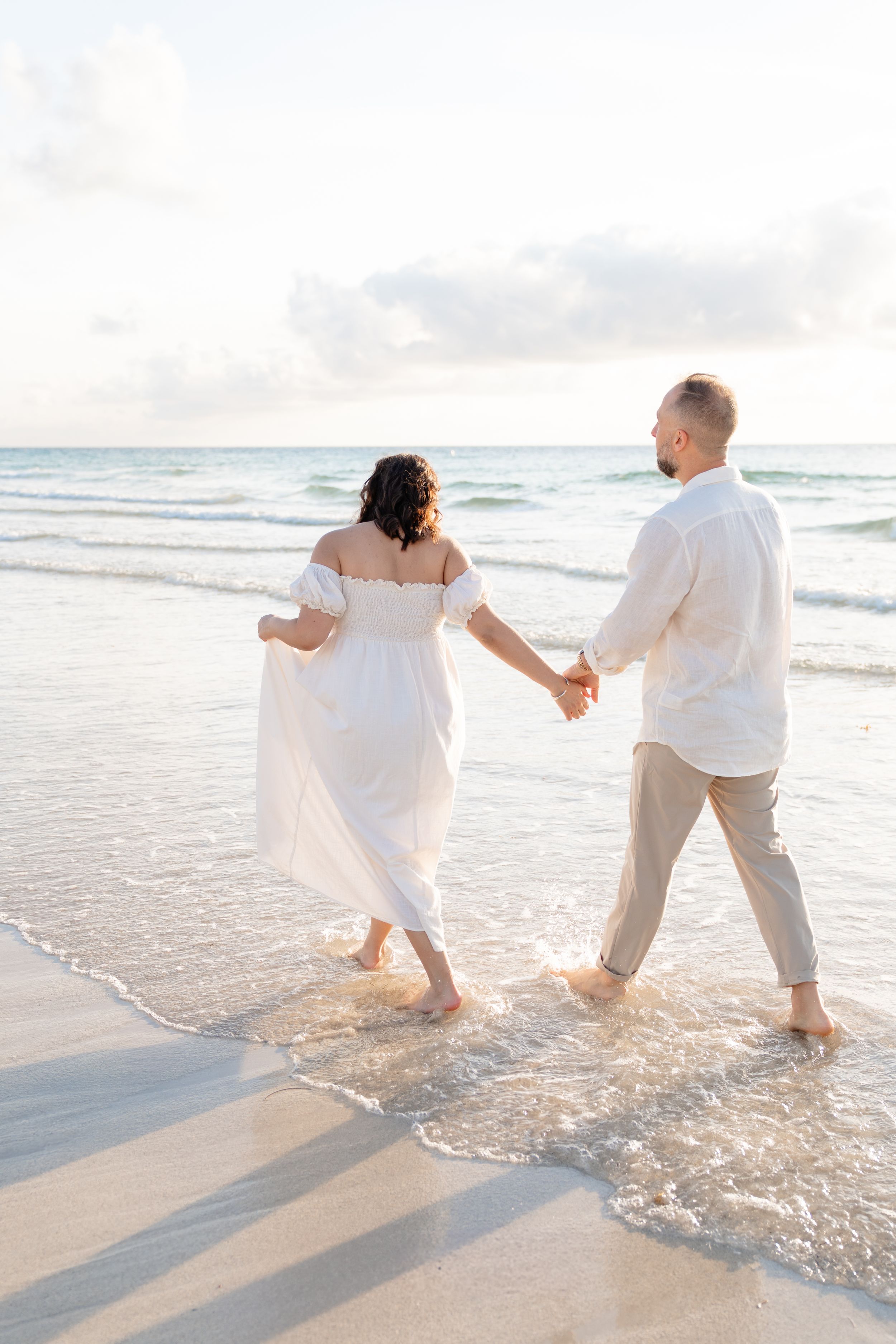 A couple in white clothing holds hands while splashing along the shoreline at sunset creating playful beach photos.