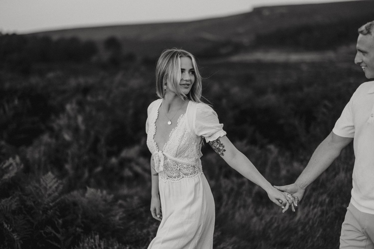 Black and white photo of couple holding hands while walking in an outdoor field with hills in the background. By Lincolnshire photographers.