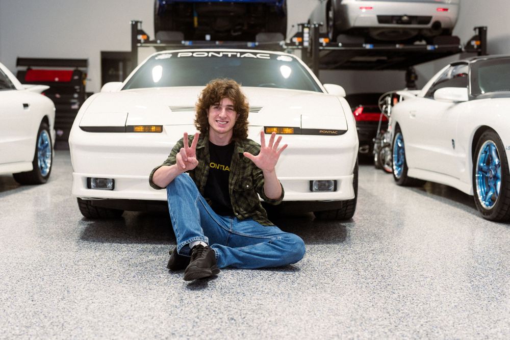 Automotive enthusiast posing between white sports cars in a garage with a blue vehicle on a lift.