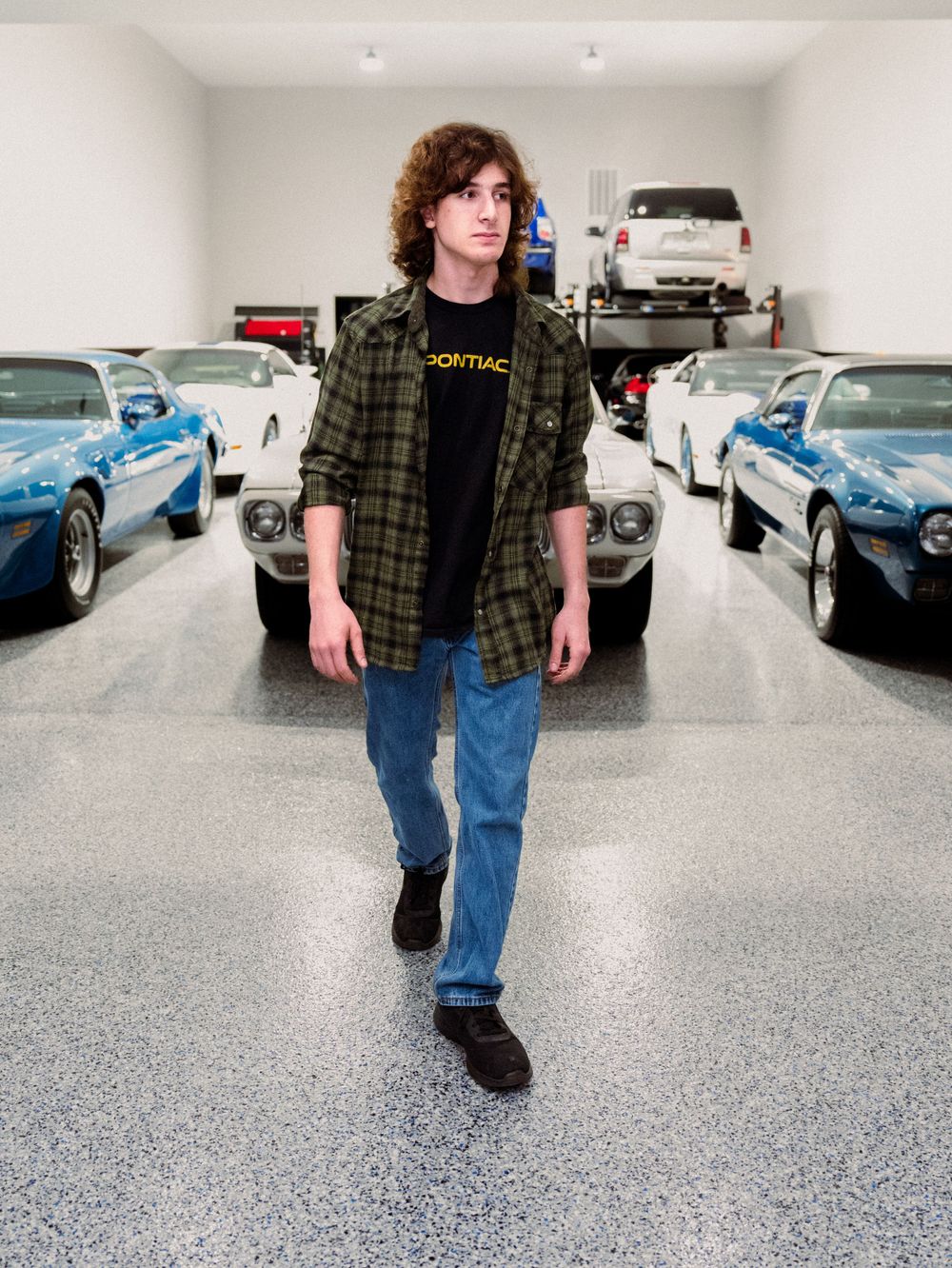 Person wearing plaid shirt and jeans poses in front of vintage cars in garage.