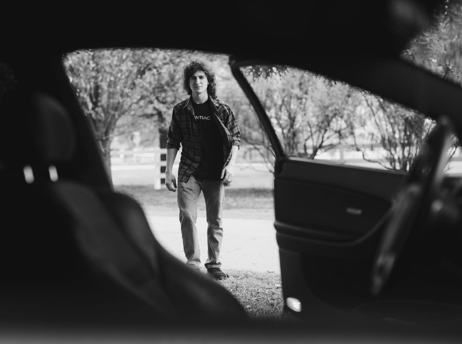 Black and white view through car window of person standing on tree-lined path.