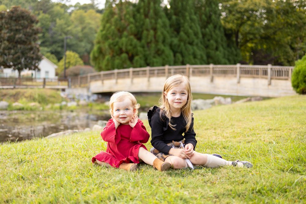 Two children sitting together on grass near a wooden bridge in a park setting.