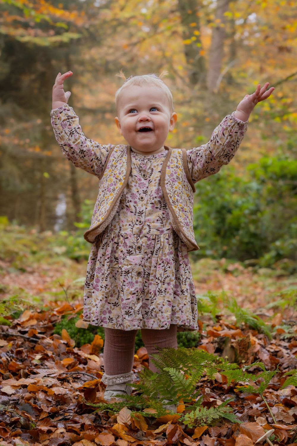 Toddler in floral dress raises arms joyfully among fallen autumn leaves.
