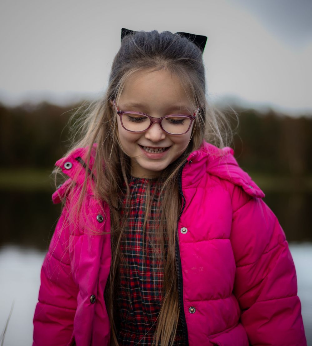 Young person in bright pink winter coat and glasses smiles warmly.