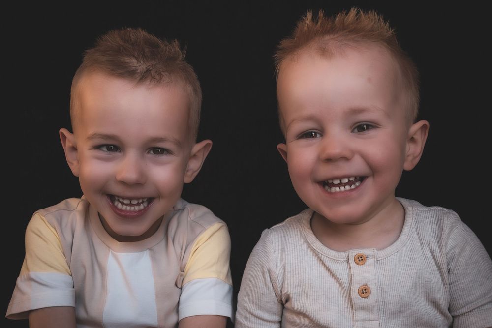 Two smiling young children sitting together in white shirts with similar spiky hairstyles.