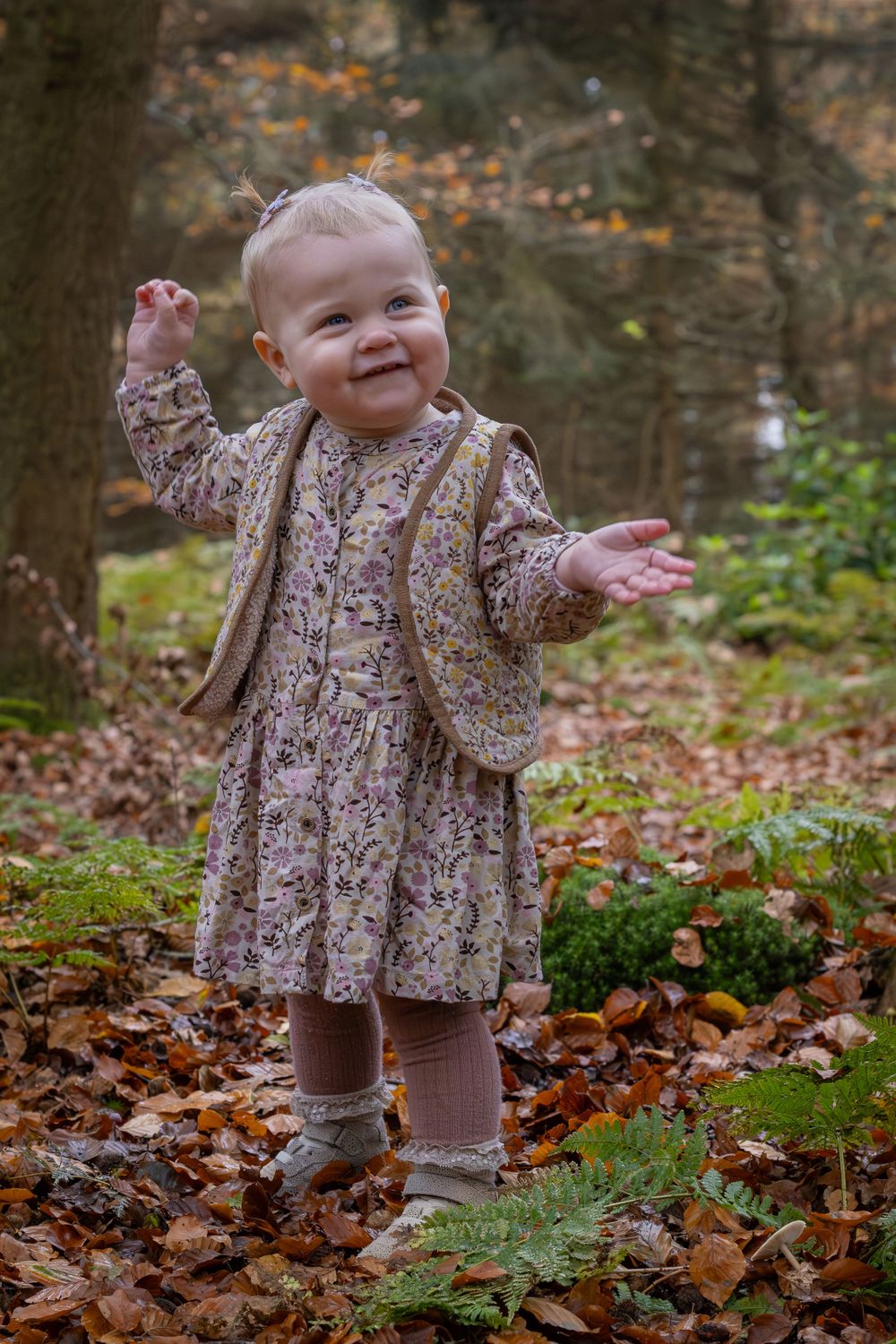 Smiling baby in floral dress playing among fallen autumn leaves in forest setting.