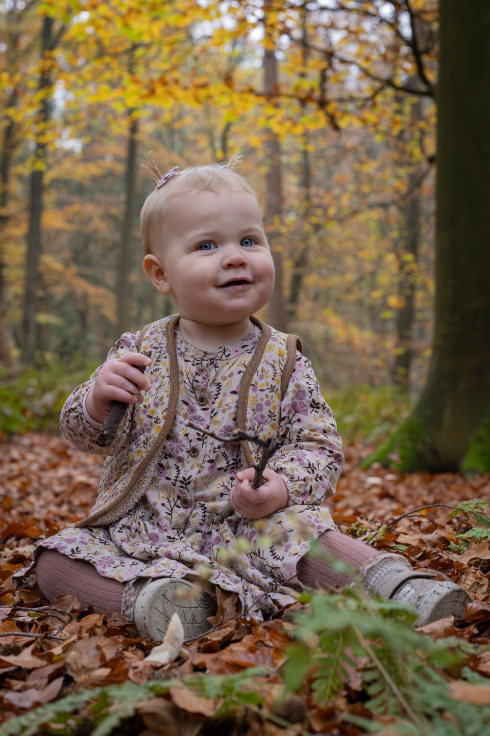 Baby in floral dress sits among fallen autumn leaves in a forest.