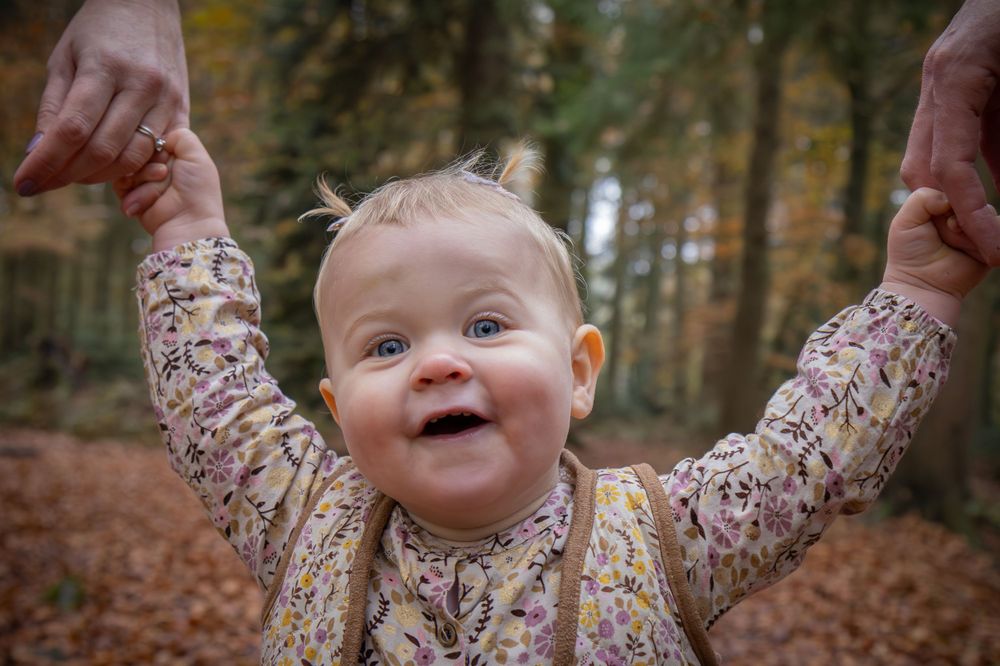 Excited toddler in floral dress raises arms with joy in autumn forest.
