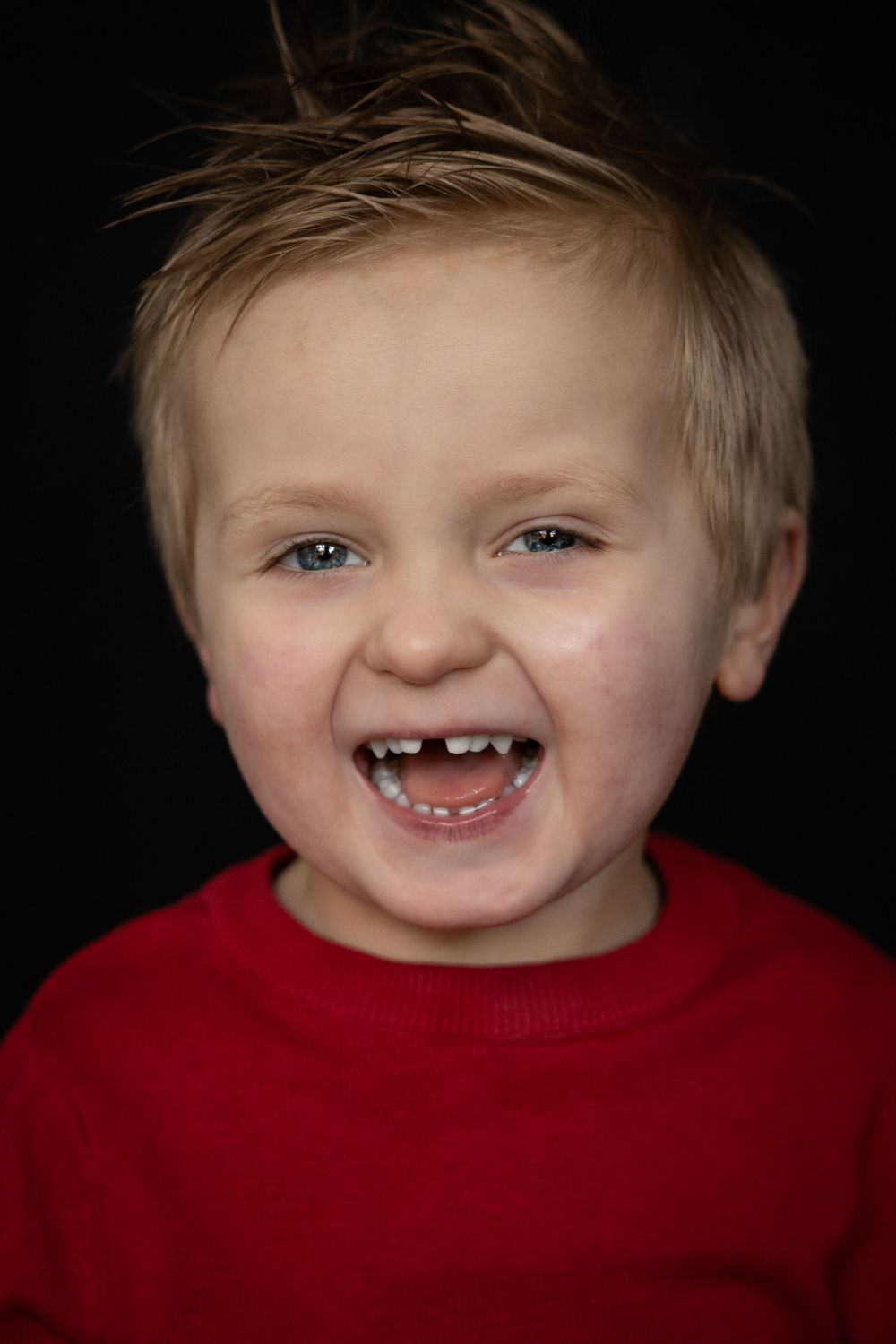 A young child with blonde spiky hair in a red sweater laughs joyfully on a black background.