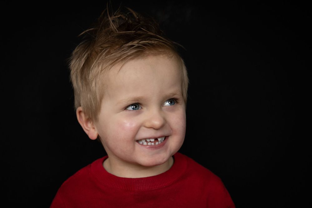Child wearing a red sweater shown smiling against a black background in a studio portrait.