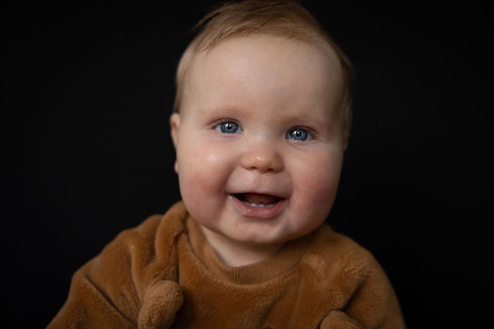 Smiling baby wearing a brown sweater photographed against a dark background.