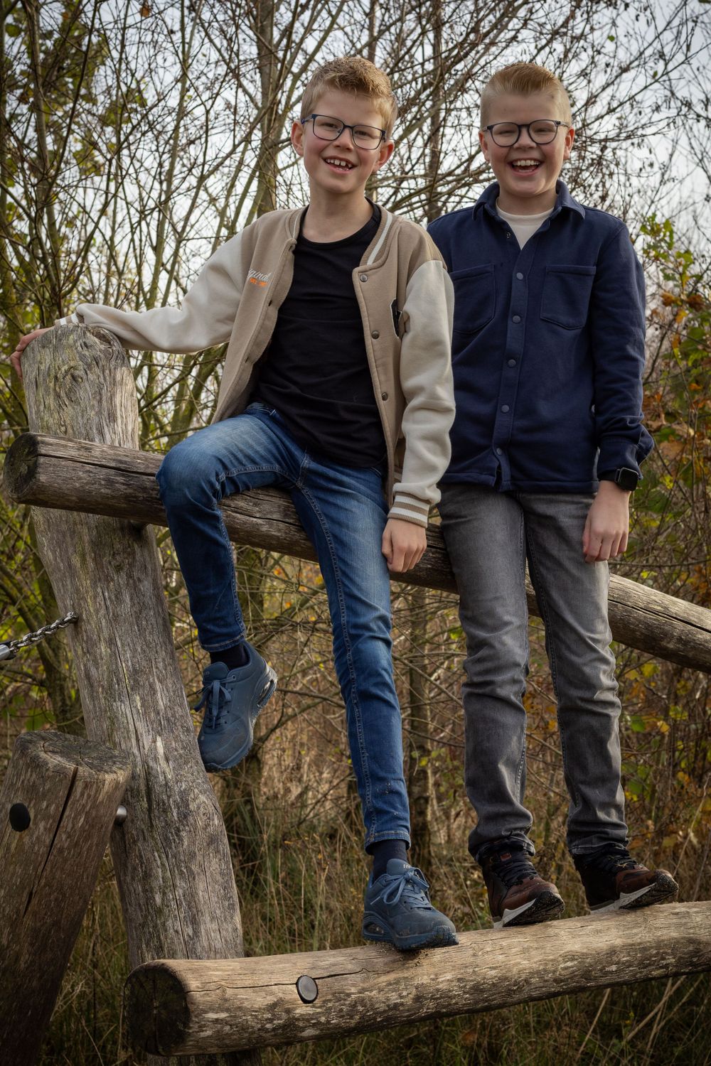 Two figures in casual wear pose on rustic wooden log against autumn foliage backdrop.