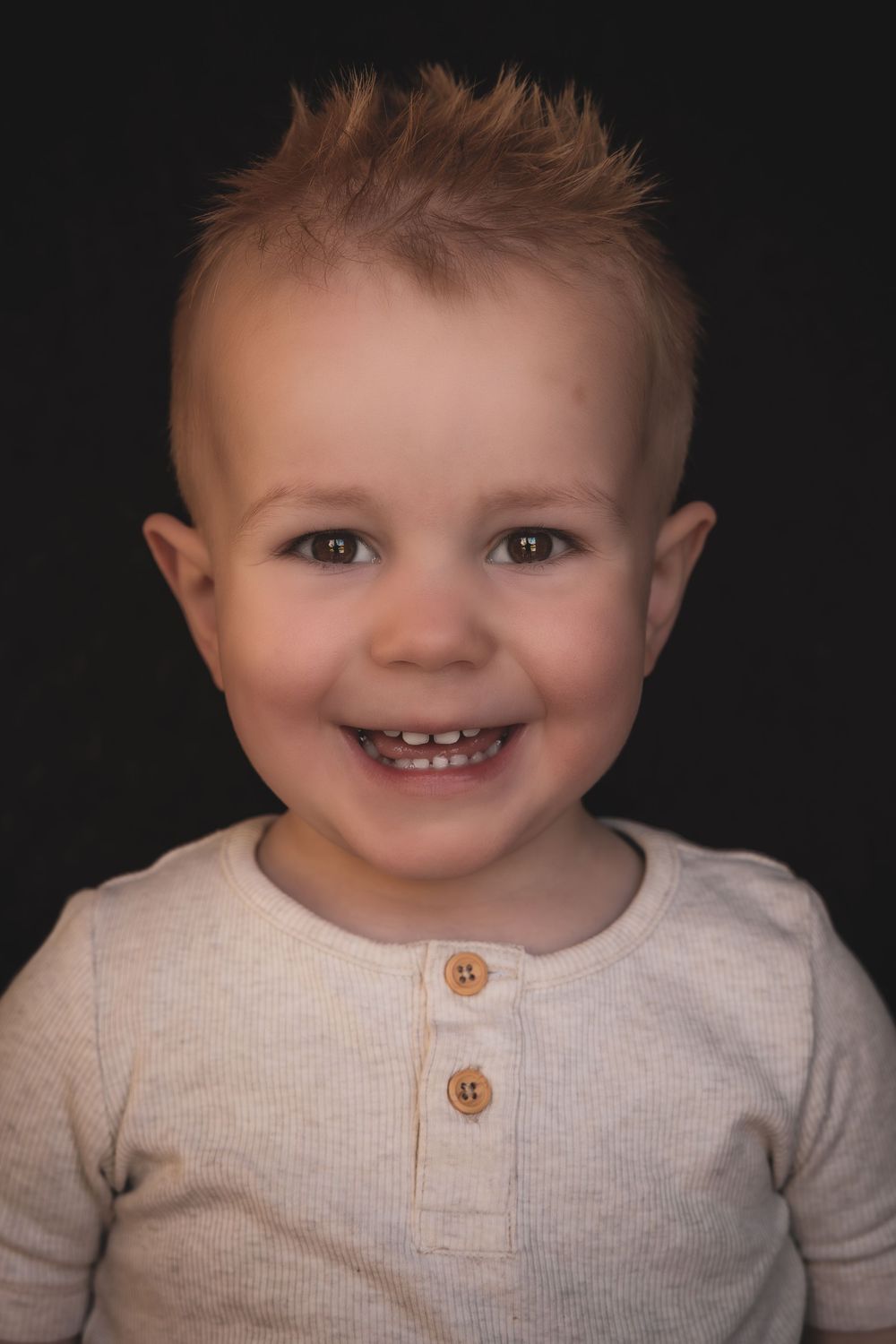 A small child wearing a beige henley shirt smiles brightly against a dark background in a studio portrait.