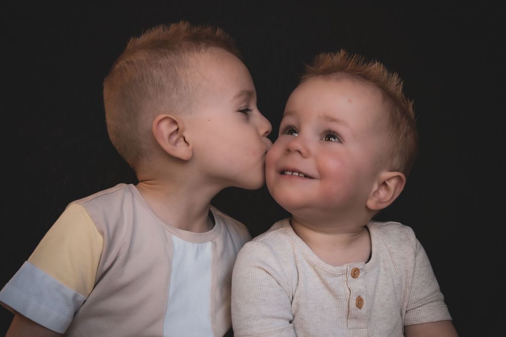 Two toddlers in light colored shirts share a sweet moment against a dark background.