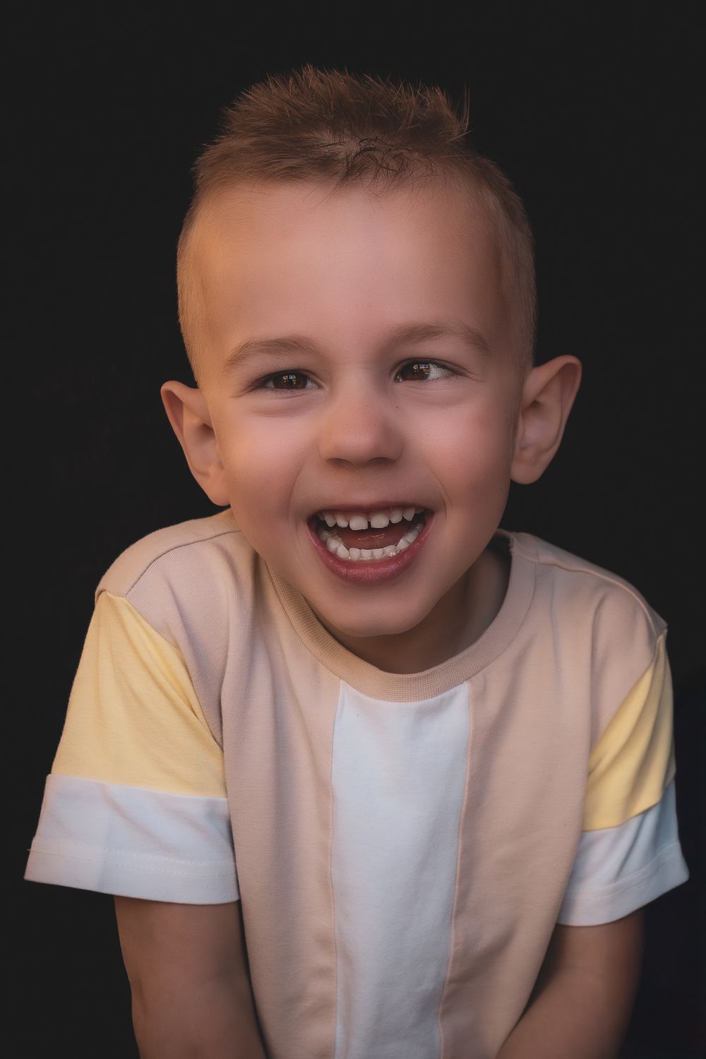 Young child wearing a white and yellow striped t-shirt smiles brightly against a dark background.