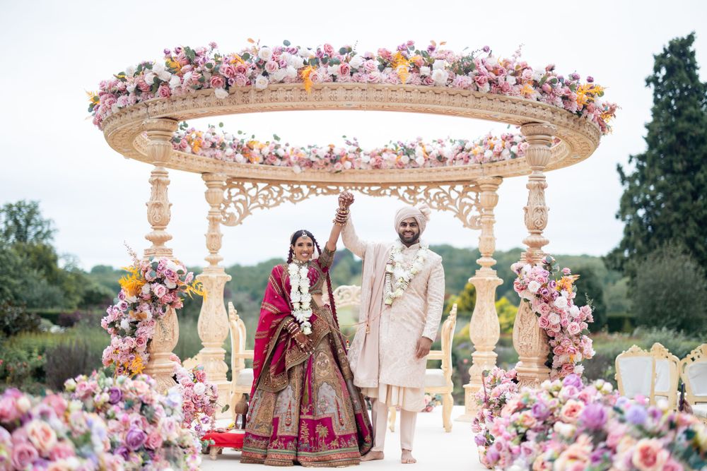 A beautiful outdoor wedding ceremony under an ornate floral mandap decorated with pink and purple blooms.