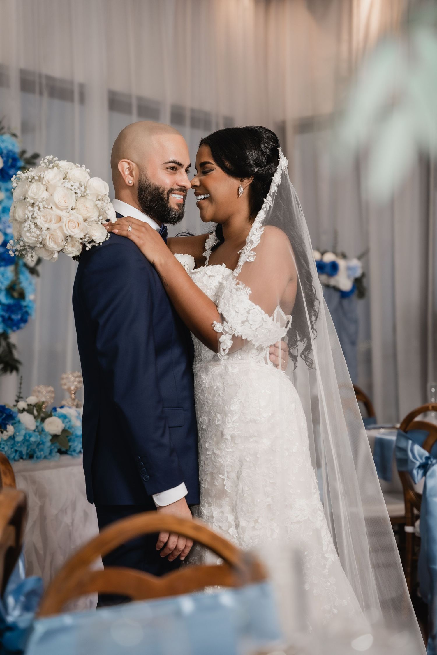 Beautiful wedding moment captured in front of draped white curtain with blue floral decor and a stunning white bouquet.