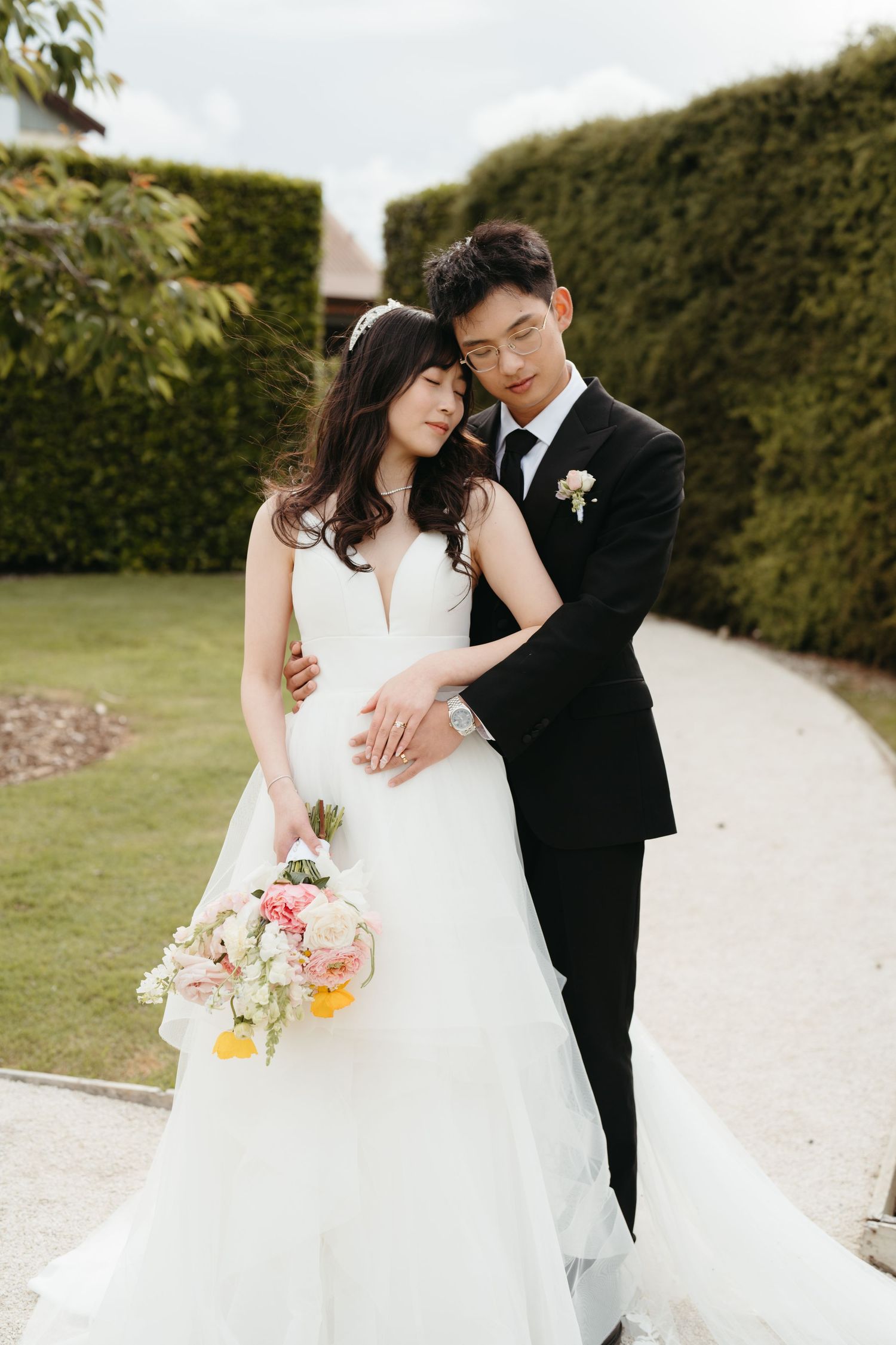 A couple in wedding attire embrace on a garden path with hedge walls, the bride holding a pastel floral bouquet.