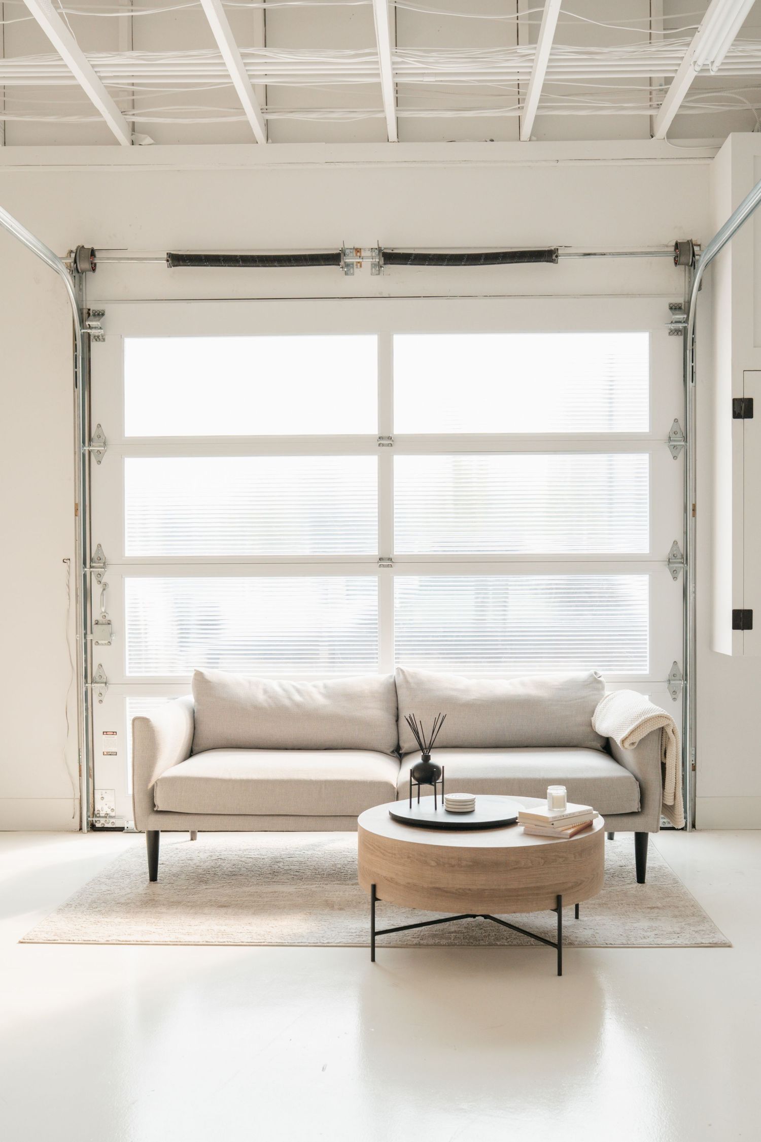 Minimalist loft space with a sleek beige sofa against white walls and industrial-style garage doors and windows.