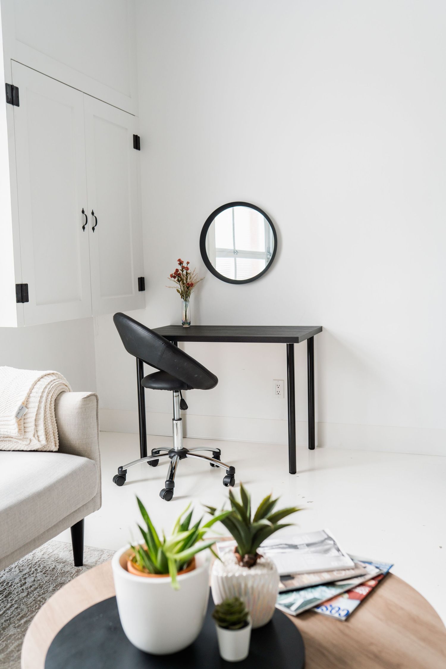 Modern minimalist workspace with black desk, round mirror, office chair, and small potted succulent plants on white walls.