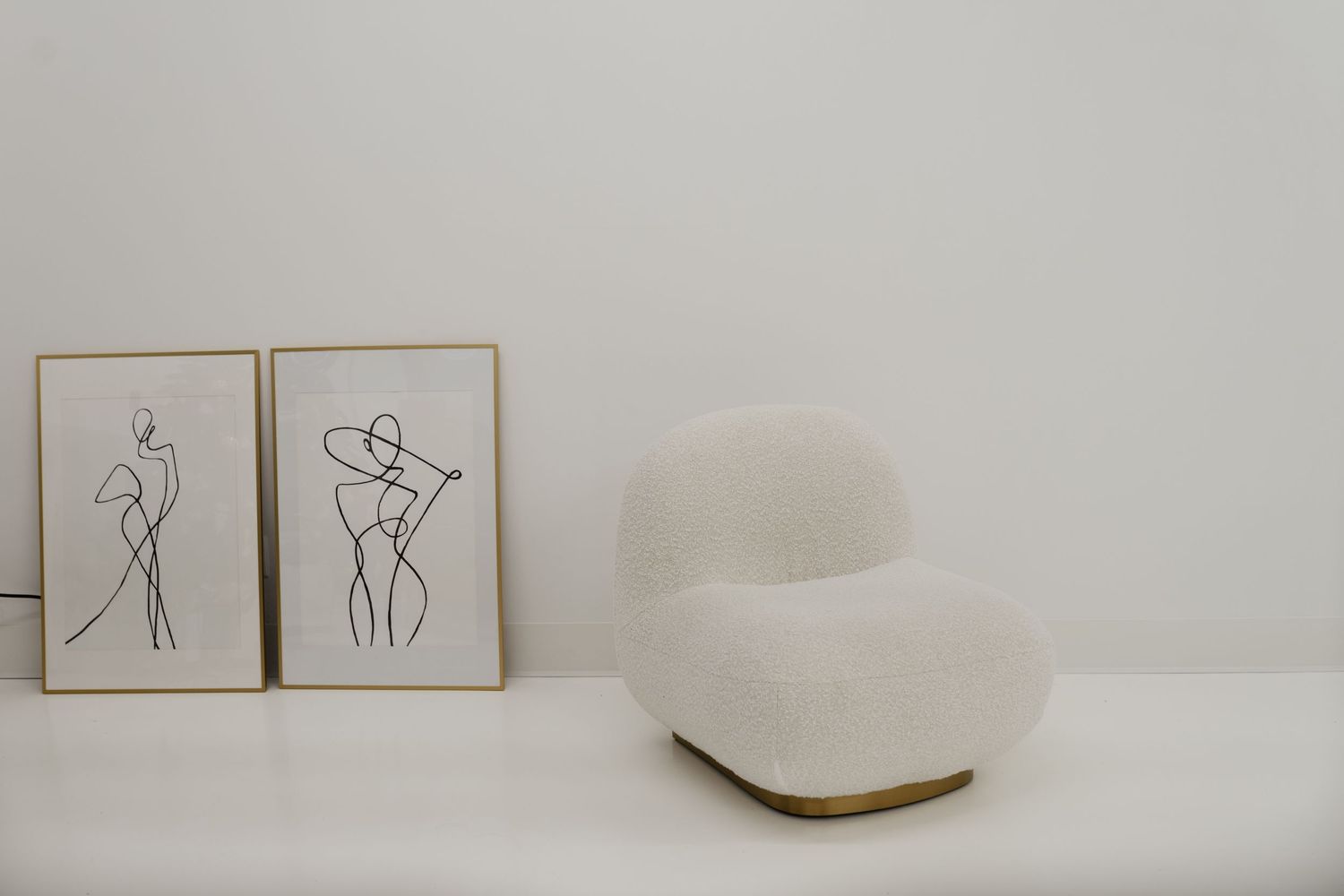 Minimalist line art drawings of figures hang above a white curved accent chair against a neutral wall.