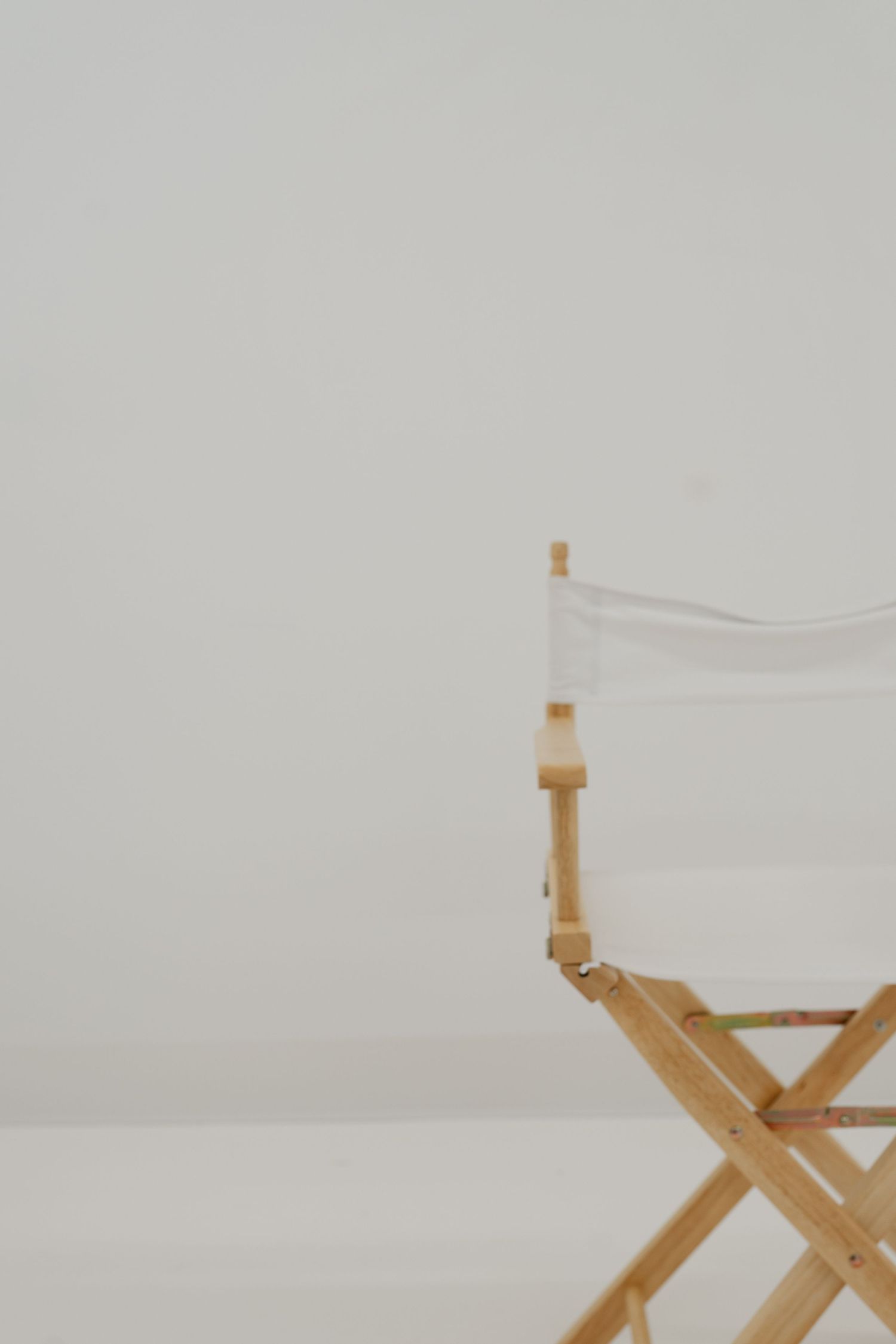 Partial view of a wooden director's chair with white canvas against a minimalist light grey background.