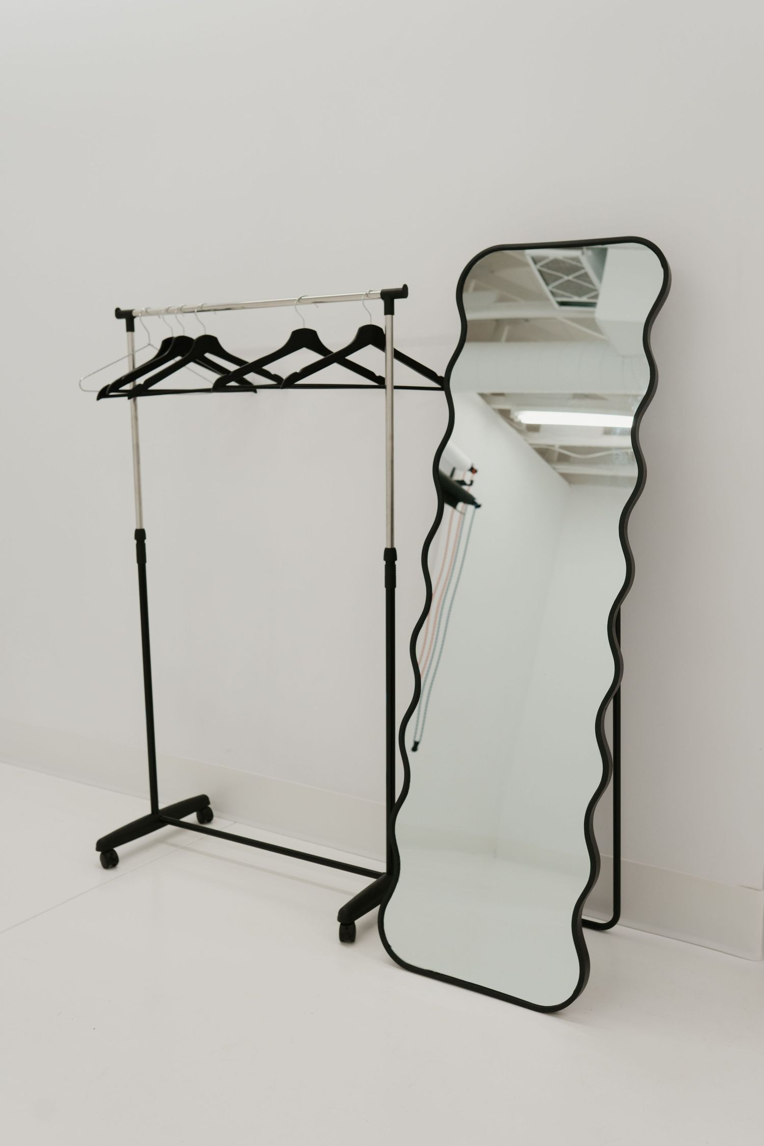 Minimalist clothing rack with black hangers and a wavy-framed full-length mirror against white wall.