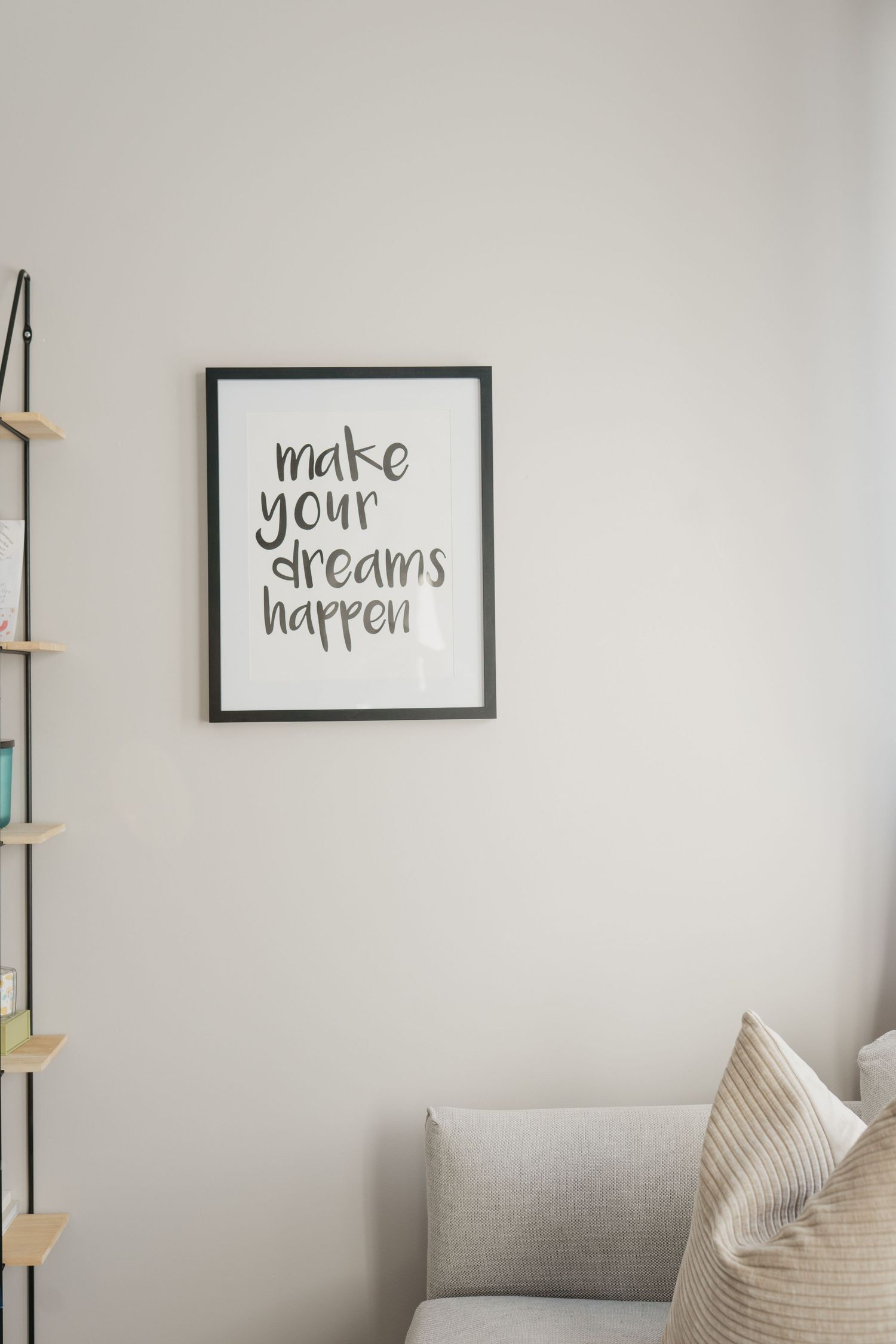 Minimalist room interior with framed motivational 'make your dreams happen' text and partial view of gray sofa.