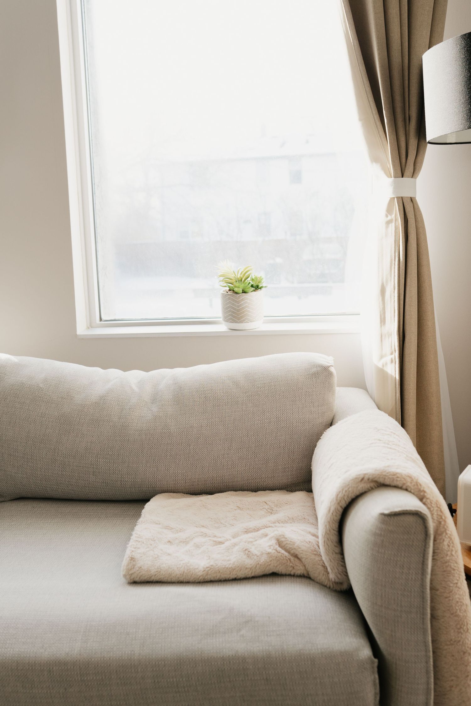 A cozy gray sofa with throw blanket by window featuring beige curtains and small potted plant on windowsill.