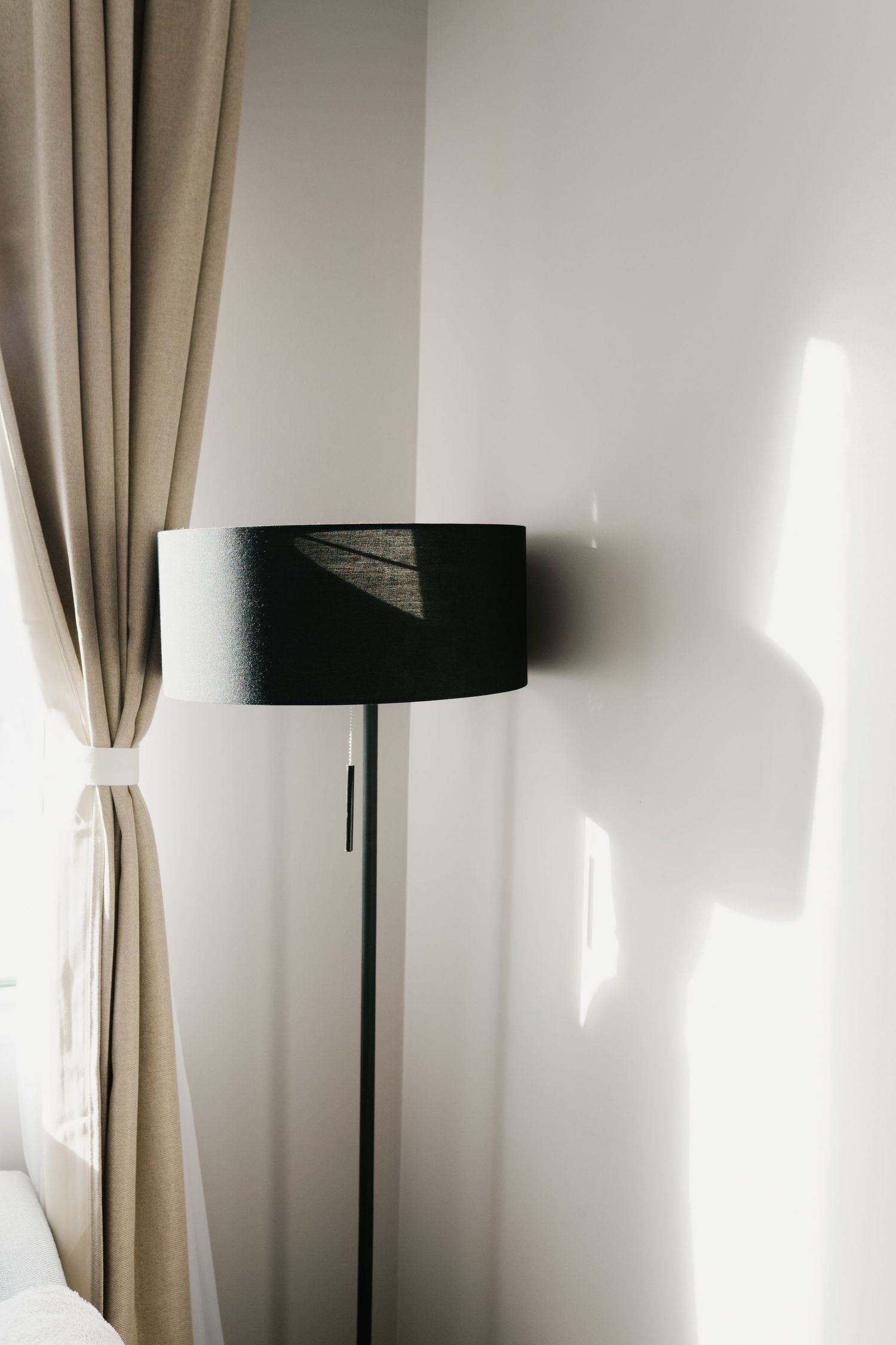 A modern black floor lamp with a cylindrical shade stands against a white wall next to beige curtains.