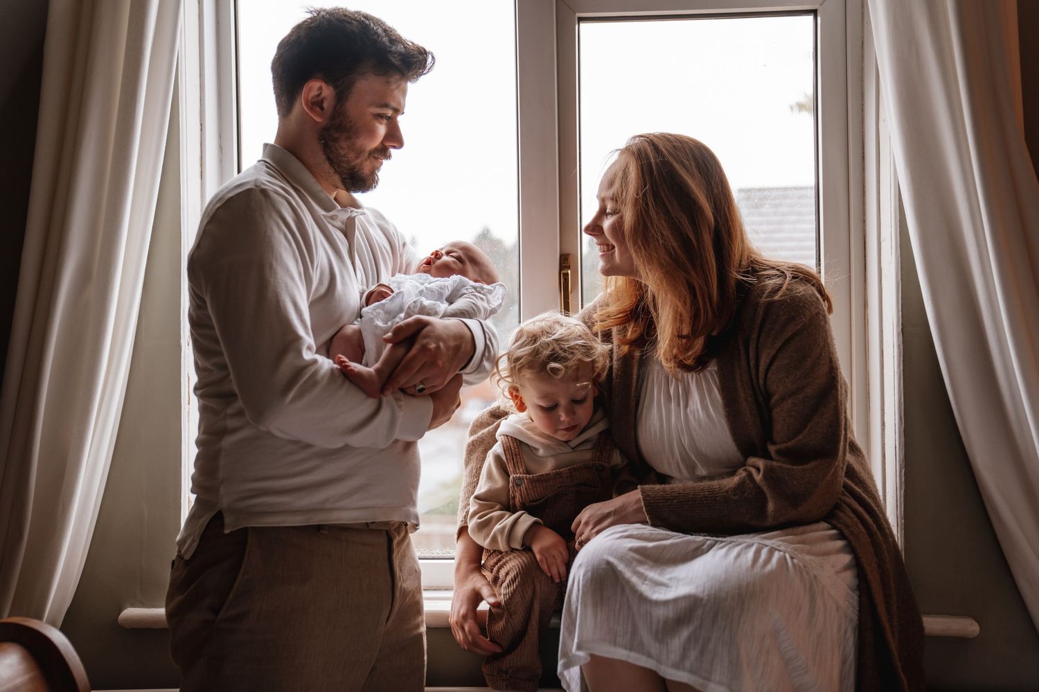 New parents smile lovingly at their newborn baby while toddler sits nearby by a bright window.
