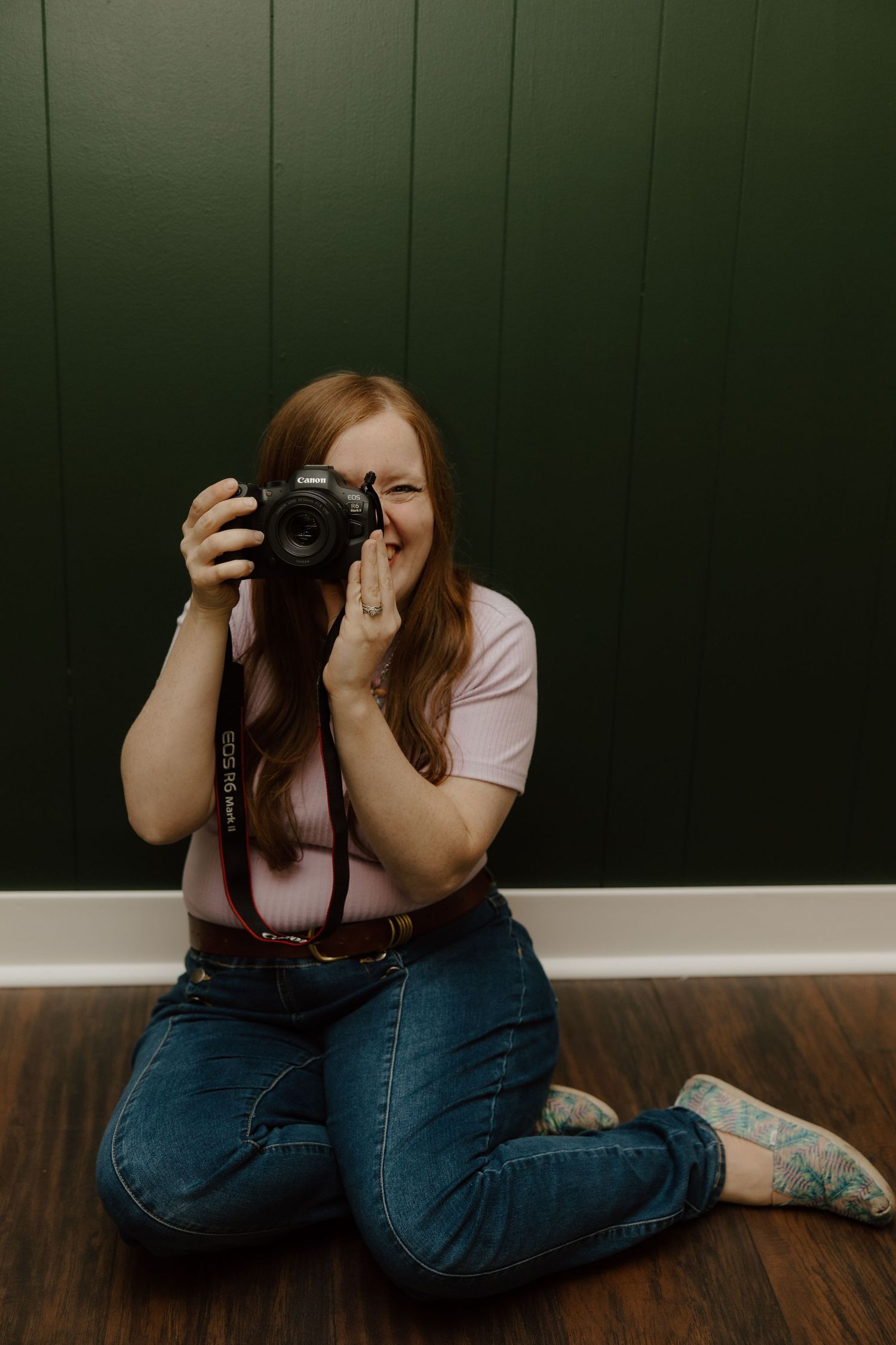 Photographer in jeans and pink shirt sits on hardwood floor taking photos against dark green wall.