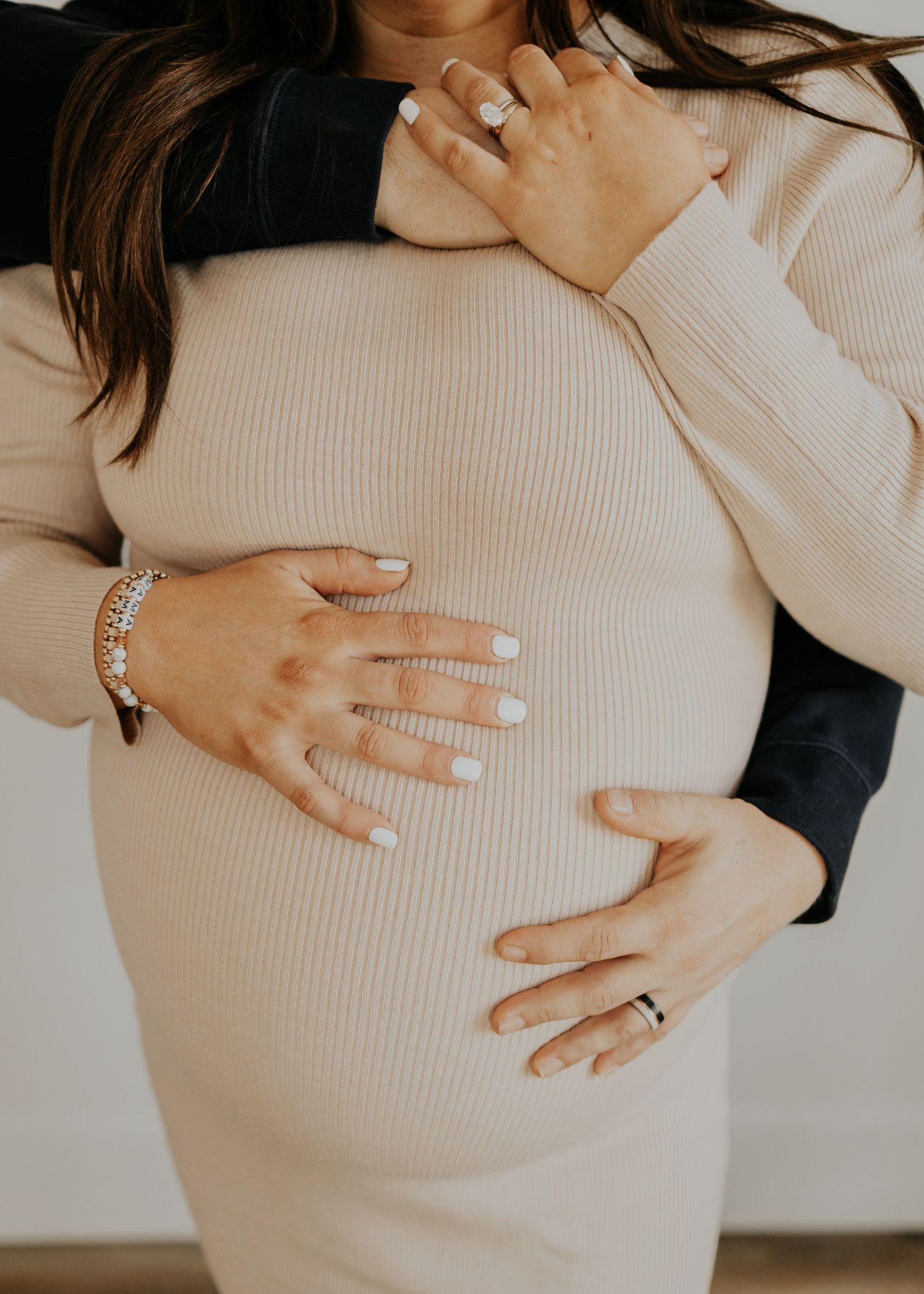 Close-up photo of hands resting on a pregnant belly wearing a cream colored ribbed sweater and delicate jewelry.