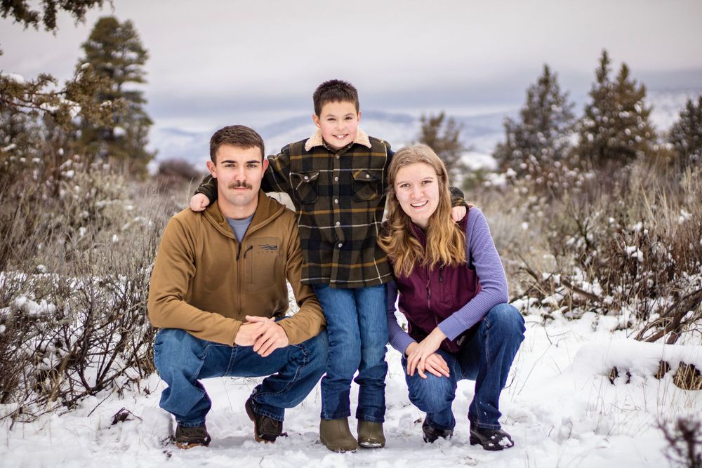 a young man and woman squat on the ground by younger brother who stands between them and puts his arms on their shoulders. there is snow