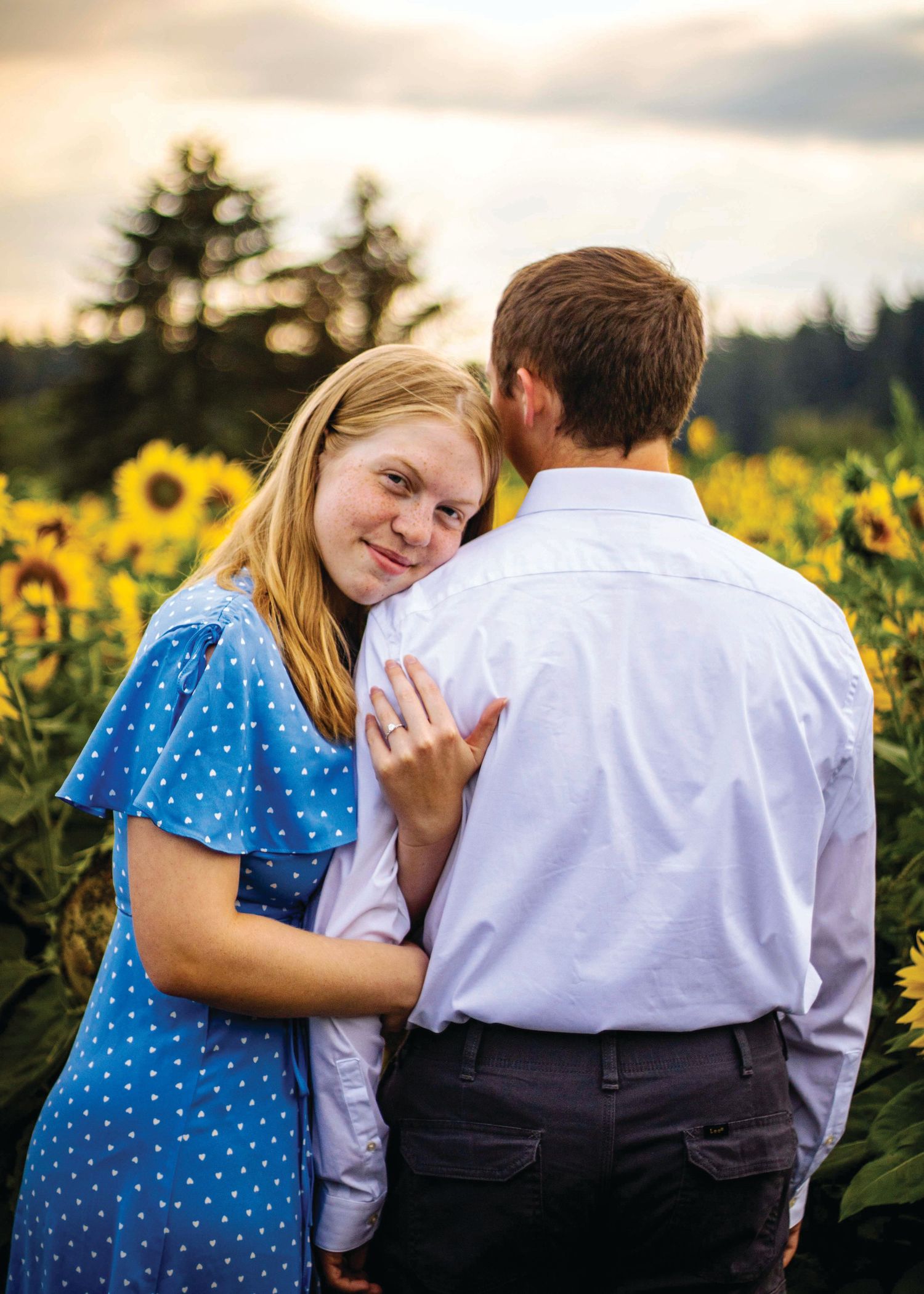 man faces away from camera, woman rests her head on his shoulder and looks at camera. they are in a field of sunflowers and she has a ring
