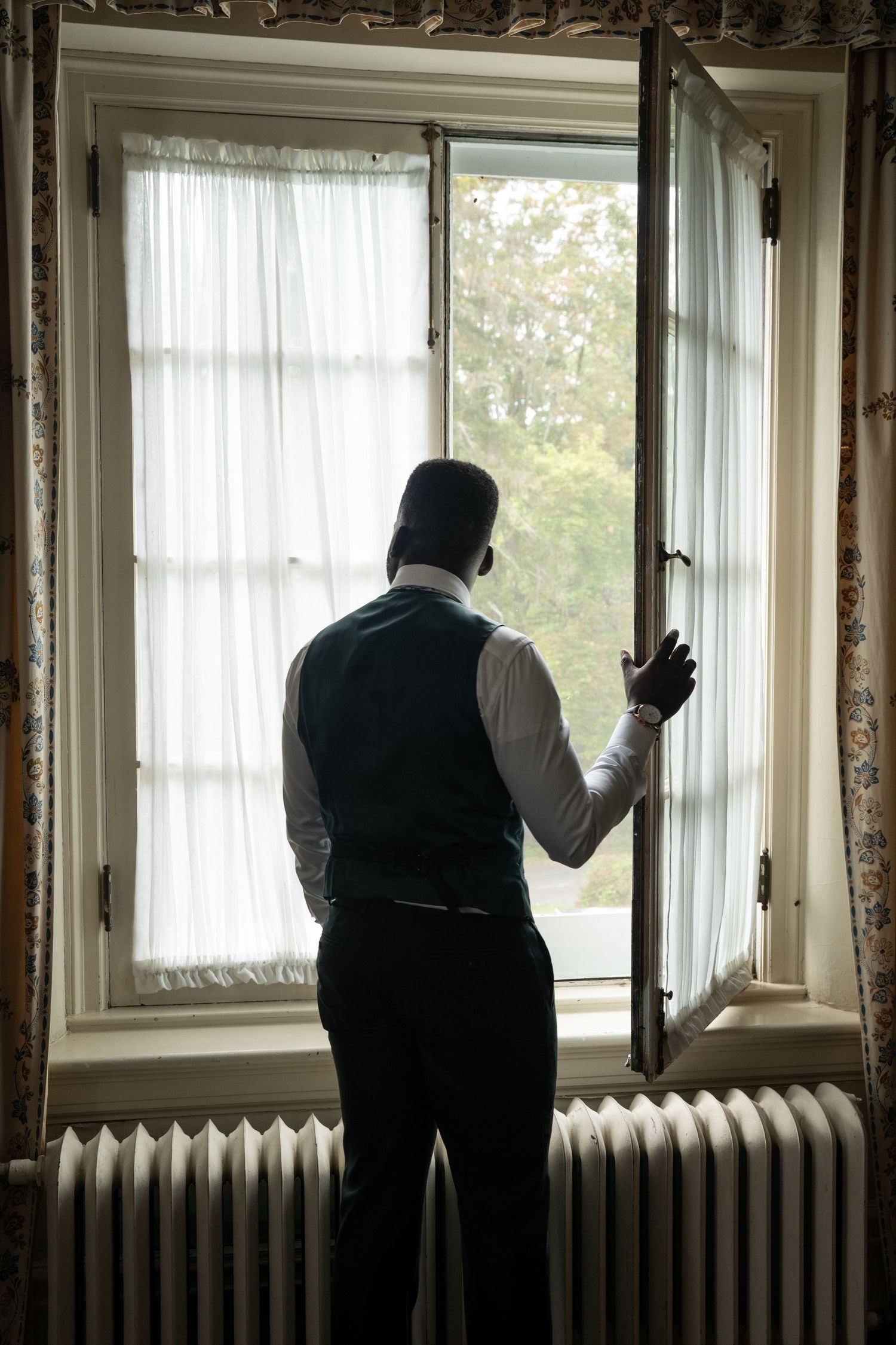 Groom in vest stands silhouetted at an open window, gazing outside of Strong Mansion on his wedding day.