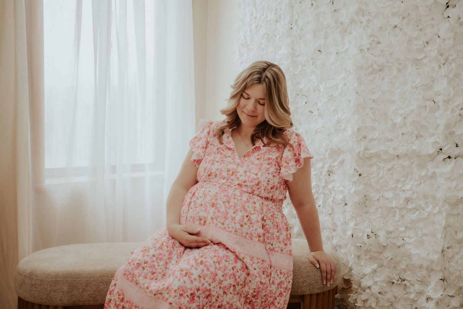 A woman in a pink floral dress sits by a window during a maternity photoshoot.
