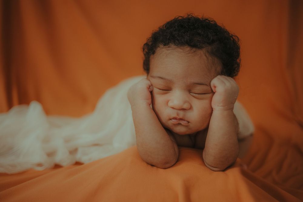 Peaceful sleeping baby resting head on hands against orange backdrop.