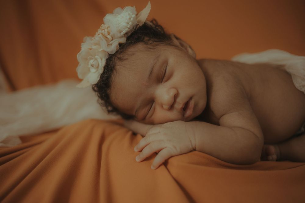 Sleeping infant wearing delicate floral crown on orange textured bedding.