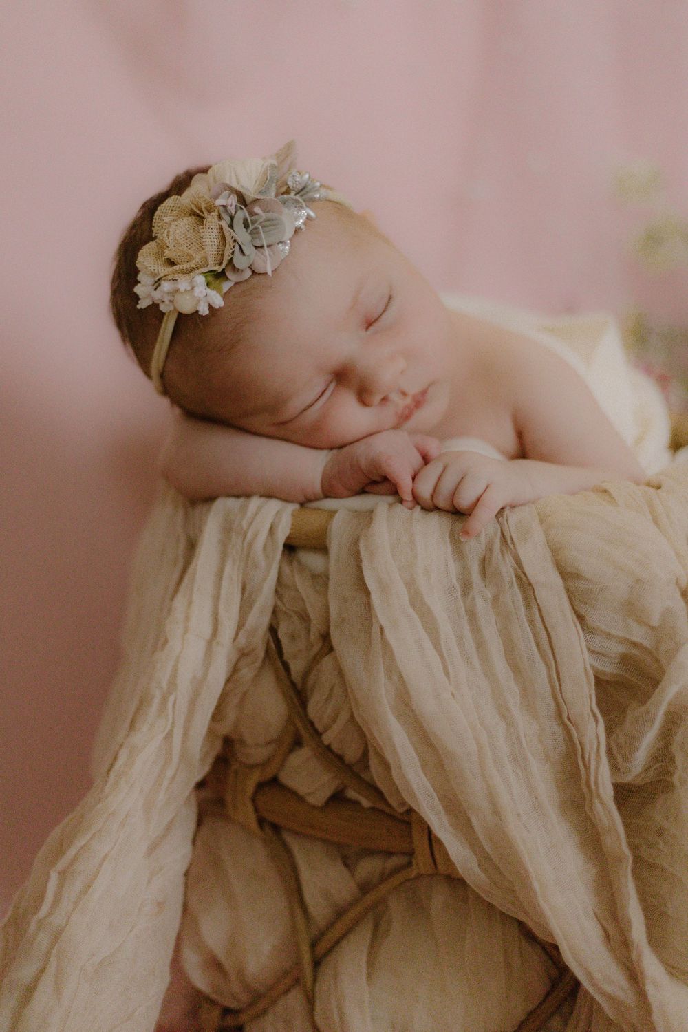 Sleeping newborn resting peacefully on cream colored fabric wearing delicate floral headband in soft lighting.