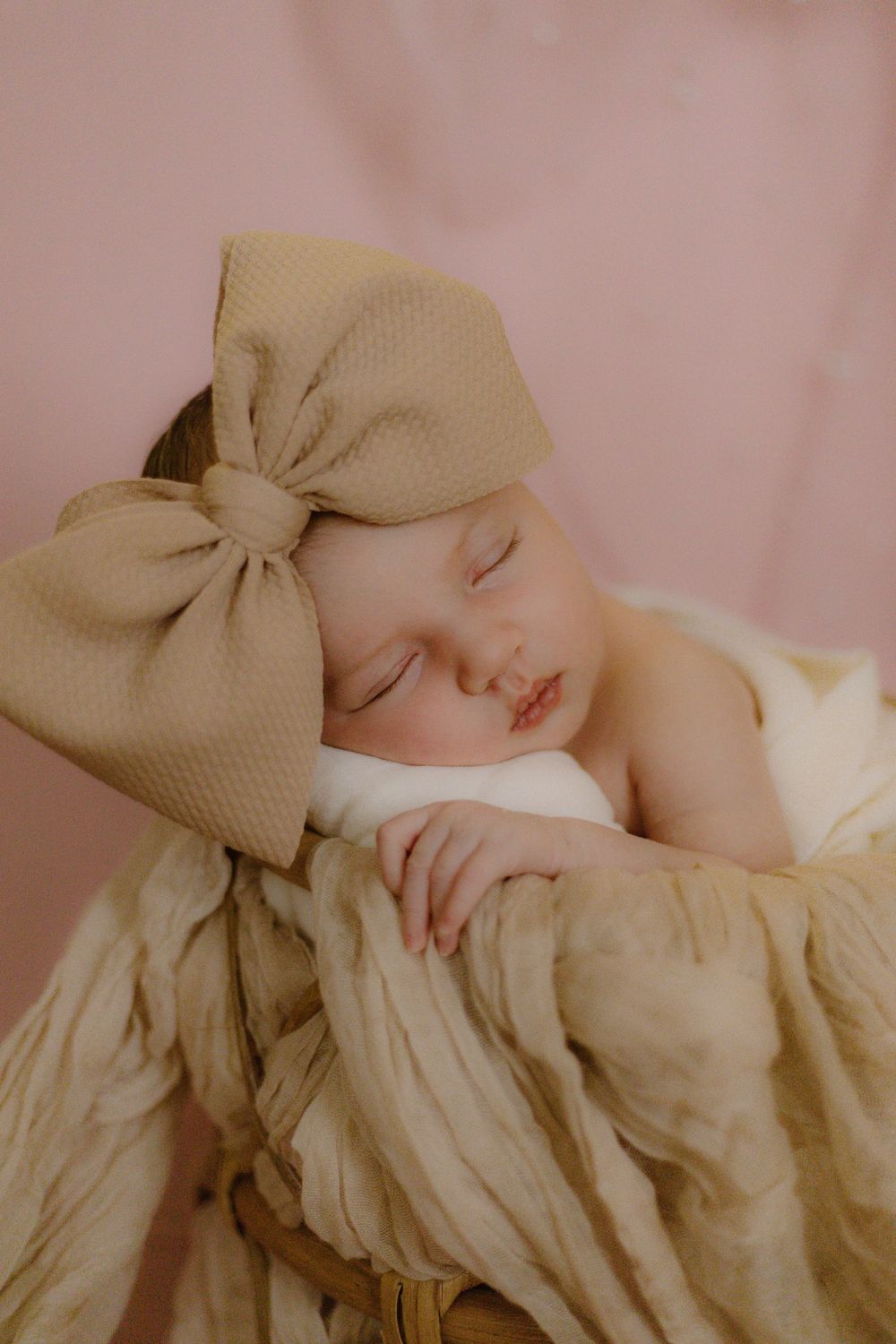 Sleeping newborn wearing a large beige bow headband resting on cream-colored textured fabric with pink background.